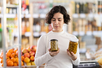 In store, woman near counter looking at pickled green olives jar. Visitors make choice of...