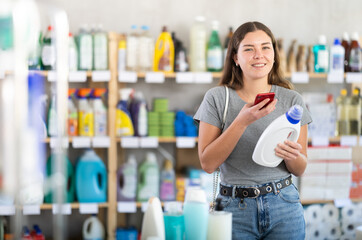 Interested smiling young girl using mobile app to scan barcode on bottle of detergent for discounts...