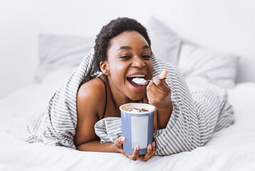 Eating in bed. Happy african american girl puts spoon in her mouth and holds bucket of ice cream at...