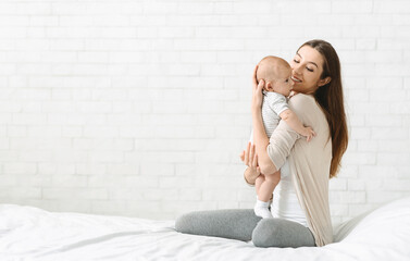 Happy young woman cuddling her newborn child while sitting on bed at home, enjoying her maternity...