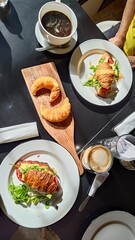 Top-down view of a delicious french breakfast spread on a dark table. Features croissant sandwiches with fresh greens, plain croissants on a wooden board, and two cups of coffee under bright sunlight.
