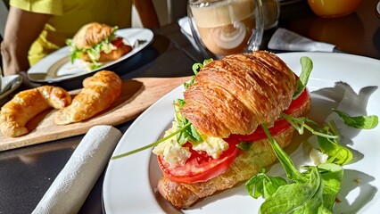 Delicious croissant sandwich with scrambled eggs, fresh tomato, and arugula on a white plate. A perfect breakfast or brunch meal served with a cup of coffee in a cozy cafe setting. Healthy and tasty.