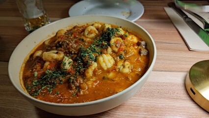 Close-up of a rustic white bowl filled with delicious potato gnocchi in a hearty tomato and meat ragu sauce, garnished with fresh parsley. Served on a wooden table in a very cozy restaurant setting.