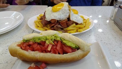 Close-up of a Chilean hot dog, known as a Completo, with tomato, avocado, and mayo. In the background, a plate of Chorrillana with fries, beef, sausage, and fried eggs. South American food.