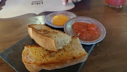 A traditional Spanish appetizer, pan con tomate, with toasted artisan bread served on a rustic slate platter with sides of tomato spread and yellow garlic aioli in a restaurant setting.