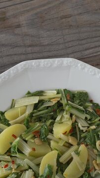 South Croatian Blitva s Krumpirom dish on a white plate. The dish is made with boiled potatoes, swiss chard, garlic, and olive oil. Rustic wooden table with copy space. Table spin, vertical video.