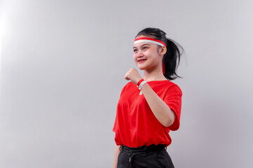 Confident young Indonesian woman wearing red shirt and red-white headband, making a fist gesture on...