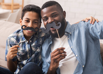 Black father and his preteen son taking selfie, holding fake moustache on sticks and smiling at...