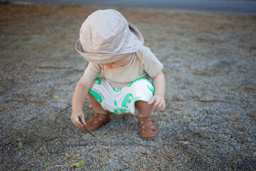 Cute toddler boy in summer clothes and sun hat squatting on gravel path and exploring nature with a twig during outdoor play on a sunny day