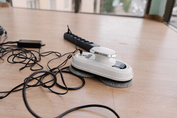 Window cleaning robot resting on floor, plugged into power strip, charging before window-washing operation at home, nobody. Modern window cleaning robot connected to chargers and power strip.