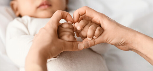 Maternal Love Concept. Closeup of young African American mom holding her cute little baby hands, making heart shape with fingers. Child wearing bodysuit lying on the white blanket sheets at home