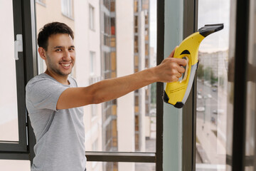 Portrait of friendly male cleaning service employee washing window glass with cordless vacuum cleaner, ensuring spotless and shiny finish in modern apartment interior. Concept of maintenance appliance