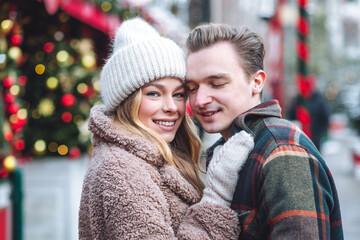 Fototapeta premium Beautiful young loving couple, boyfriend and girlfriend having fun on a christmas market wearing warm clothes. Outdoors, winter time, snowy weather. Tangerines in string bag, fir tree branches