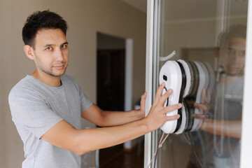 Portrait of handsome young man installing robotic vacuum cleaner on window, preparing to activate device for efficient cleaning session, ensuring spotless interior environment, looking at camera.