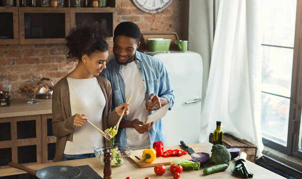Black couple using digital tablet with recipe while preparing dinner in loft kitchen