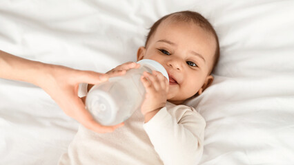 Obraz premium Nursery And Childcare Concept. Portrait of cute little black kid drinking milk, mother holding baby bottle in hands, feeding her child who lying on white blanket on the bed in the bedroom at home