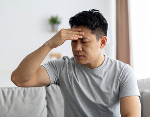 Asian Man Experiencing Headache, Touching Forehead, Eyes Closed, Indoor Setting.