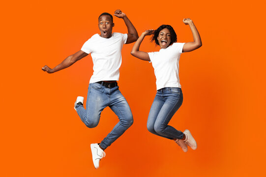 Happy black man and woman celebrating success, full length photo, copy space. Cheerful african american young couple clenching fists and screaming, jumping up over orange studio background