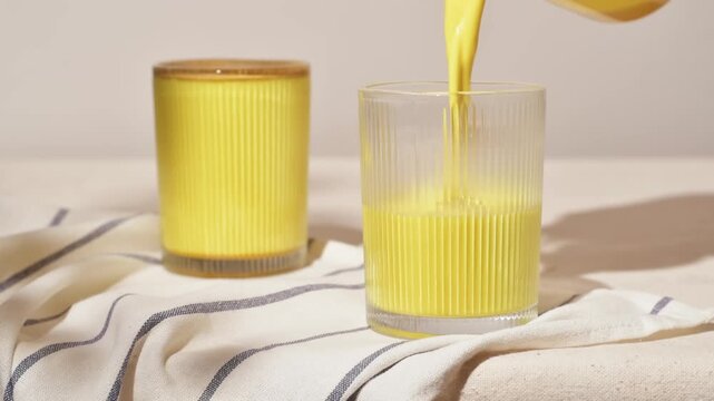 Bright golden latte pours smoothly into ribbed glass, sunlit on a striped napkin, creating cozy healthy morning atmosphere.