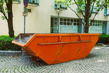 A bright orange construction dumpster is placed on the urban pavement, surrounded by lush trees and...