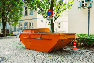 An Urban Construction Site with a bright orange dumpster contrasts with lush greenery and paved...