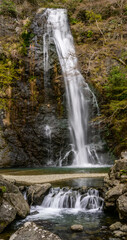 Fototapeta premium A tall waterfall in Minoh National Park, Osaka, Japan. The image has a long exposure, highlighting the water's smooth flow. Rocks and lush vegetation frame the scene, evoking a sense of tranquility.