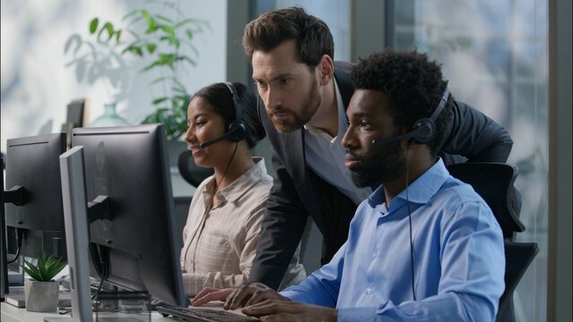 Multiethnic callcenter smiling Caucasian boss explain pointing pc monitor Indian woman employee office teamwork business call center customer support training African man headset helpline hotline desk