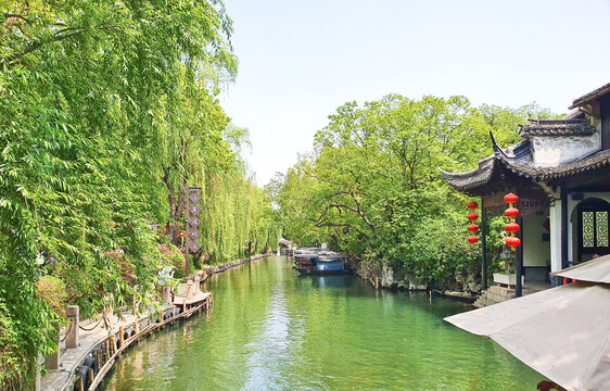 Peaceful canal scene with lush greenery, traditional Chinese architecture, red lanterns, and a walking path under clear blue sky 
