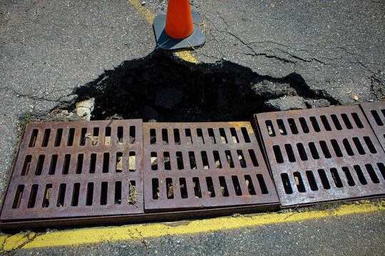 a significant pothole in asphalt pavement, located directly adjacent to a metal storm drain or catch basin grate. an orange construction cone is placed for safety. road repair season.