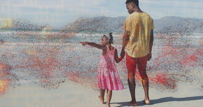 Walking father in yellow shirt and daughter in pink dress holding hands on beach, pointing ahead