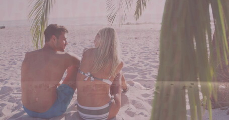 Fototapeta premium Relaxing adult couple wearing swimwear sitting on beach at sunset, with palm fronds and ocean waves