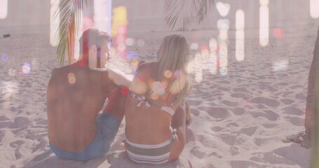 Facing ocean horizon, couple wearing swimwear sitting on sand at sunlit beach under palm fronds