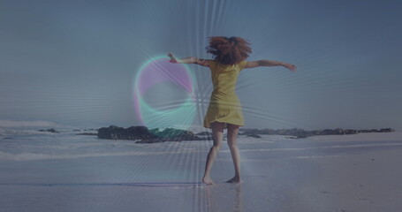 Spinning woman in yellow sundress dancing on wet sand at beach shoreline, with dark rock formations