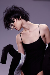 Asian woman in black bodysuit and oversized sunglasses posing with tousled short hair against gray background in stylish modern fashion atmosphere.