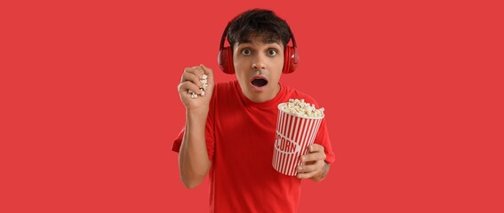 Shocked young man in headphones and with popcorn on red background