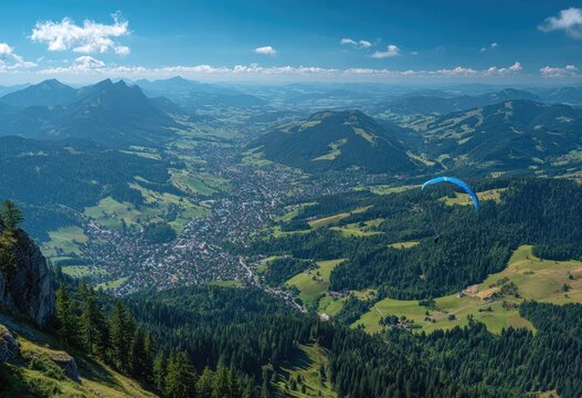 Panoramic view of a valley town nestled amongst rolling green hills and mountains under a vibrant blue sky, with a paraglider soaring overhead - Powered by Adobe