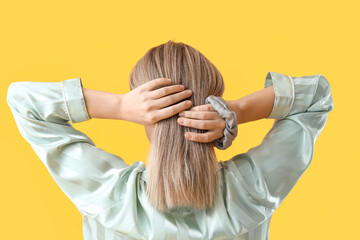 Fototapeta premium Young blonde woman in pajamas doing ponytail with scrunchy on yellow background, back view