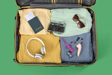 Open suitcase with clothes, sex toys, passport and accessories on green background