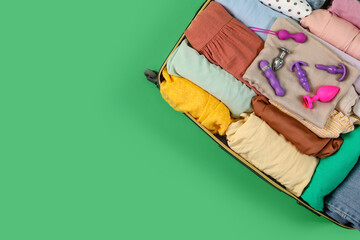 Open suitcase with clothes and different sex toys on green background