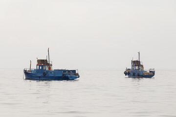 Naklejka premium Two weathered blue fishing boats rest on a calm, misty sea under overcast skies, evoking solitude and maritime simplicity.