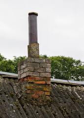 A rustic brick chimney with a metal exhaust pipe rises from a moss-covered, aging corrugated roof against an overcast sky and trees.