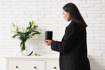 Young woman putting mortuary urn on chest of drawers near bouquet of funeral lily flowers in room