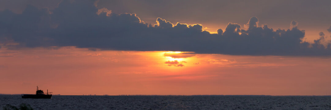 A single fishing boat drifts on Lake Ilmen as the sun sets behind dramatic clouds, casting golden and orange tones across the sky.