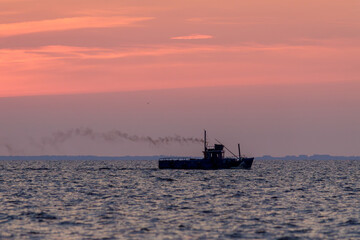 Fototapeta premium Silhouetted fishing boat releases smoke as it sails across calm ocean waters during a peaceful pink and orange sunset.