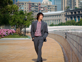 Portrait of handsome Chinese young man wearing gray suit hands in pocket walking in street, young guy with black curly hair with urban background. Male fashion, cool Asian man lifestyle.
