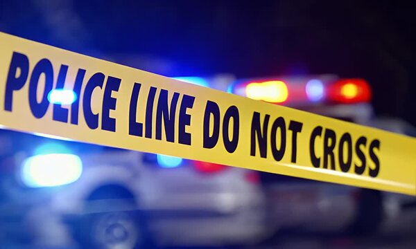 Police tape marking a restricted area at night with emergency vehicles and flashing lights in the background