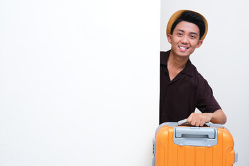 Young Asian man peeking behind wall carrying a suitcase with happy face