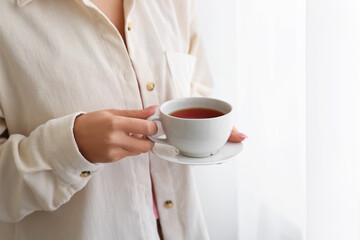 Young woman with cup of tea near light curtains at home, closeup