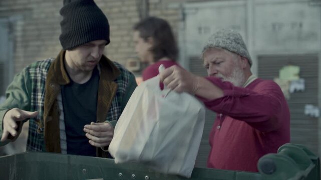Elderly homeless man and his younger friend in worn clothes examining discarded items and taking plastic bags out of garbage bin together in urban backstreet