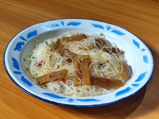 A dish of fried vermicelli with pieces of kikil, sliced chilies and vegetables, served on a blue and white patterned enamel plate on a wooden table.